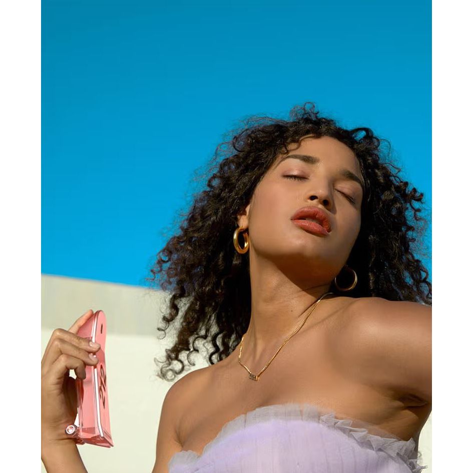 Woman with curly hair wearing makeup, holding pink cosmetic bag, gold jewelry, blue sky background