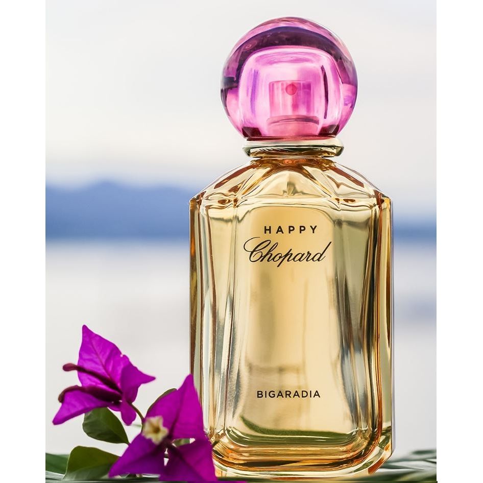 Happy Chopard Bigaradia perfume bottle with pink cap, purple flowers, and blurred background