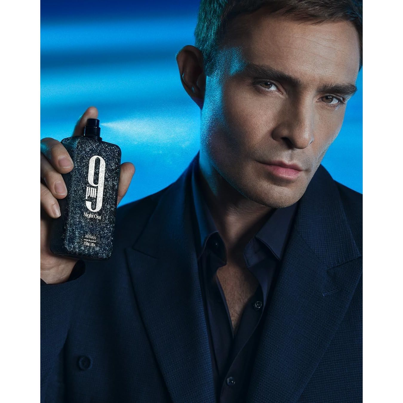 Man in navy suit holding 9pm Night Out perfume bottle against blue background